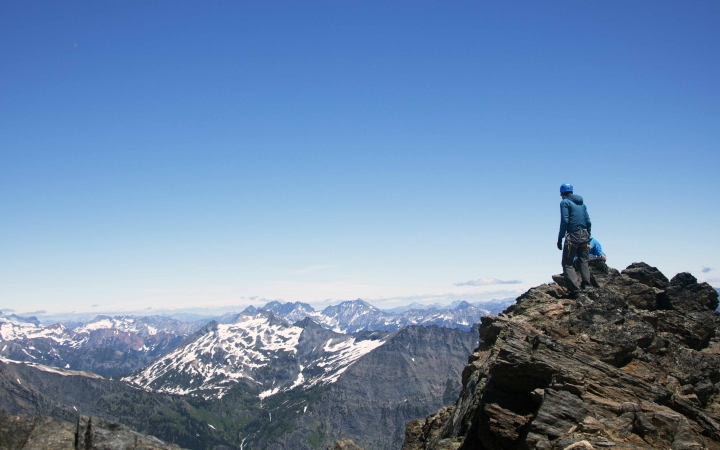 Two people stand on a rocky cliff at very high elevation, looking out over the vast mountainous and snowy landscape below.